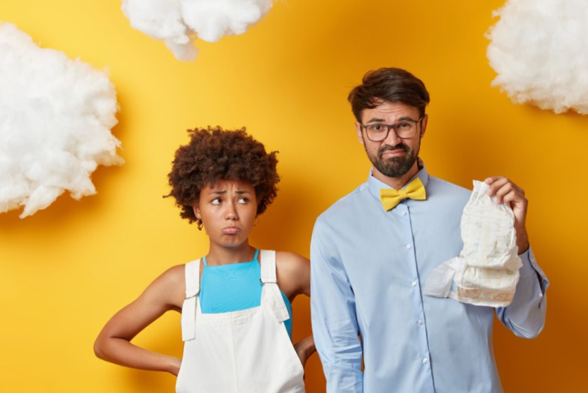 Displeased man looks with aversion at diaper being not ready to become father dressed in formal shirt. unhappy pregant woman prepares for child birth stands near husband against yellow wall