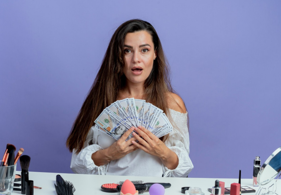 portrait of a Surprised girl holding money with makeup and beauty tools on the table  isolated on purple wall
