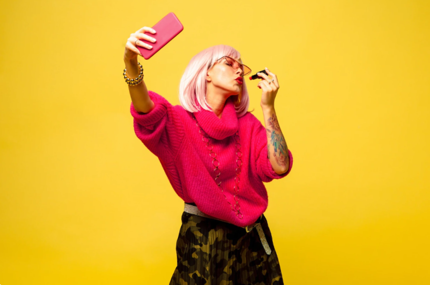 portrait on yellow background of woman taking a selfie while she's putting lipstick on her lips
