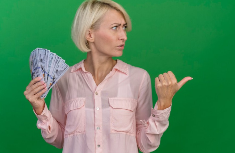 Frowning blonde woman holding money turning head to side and pointing at side isolated on green background