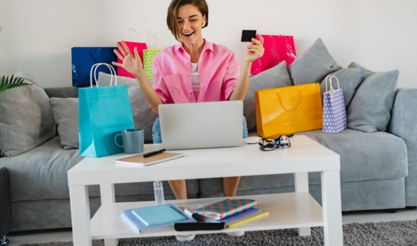happy smiling woman in pink shirt on sofa at home among colorful shopping bags holding credit card paying online on laptop
